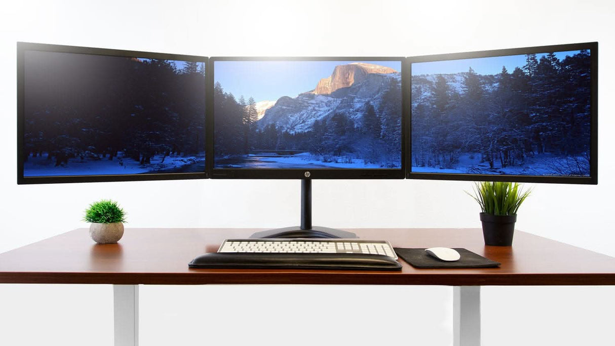 The Mount-It! Triple Monitor Stand holds three monitors displaying a mountain and forest scene, with a keyboard, mouse pad, and two potted plants on either side—all neatly arranged on a desk with tidy cables against a white background.
