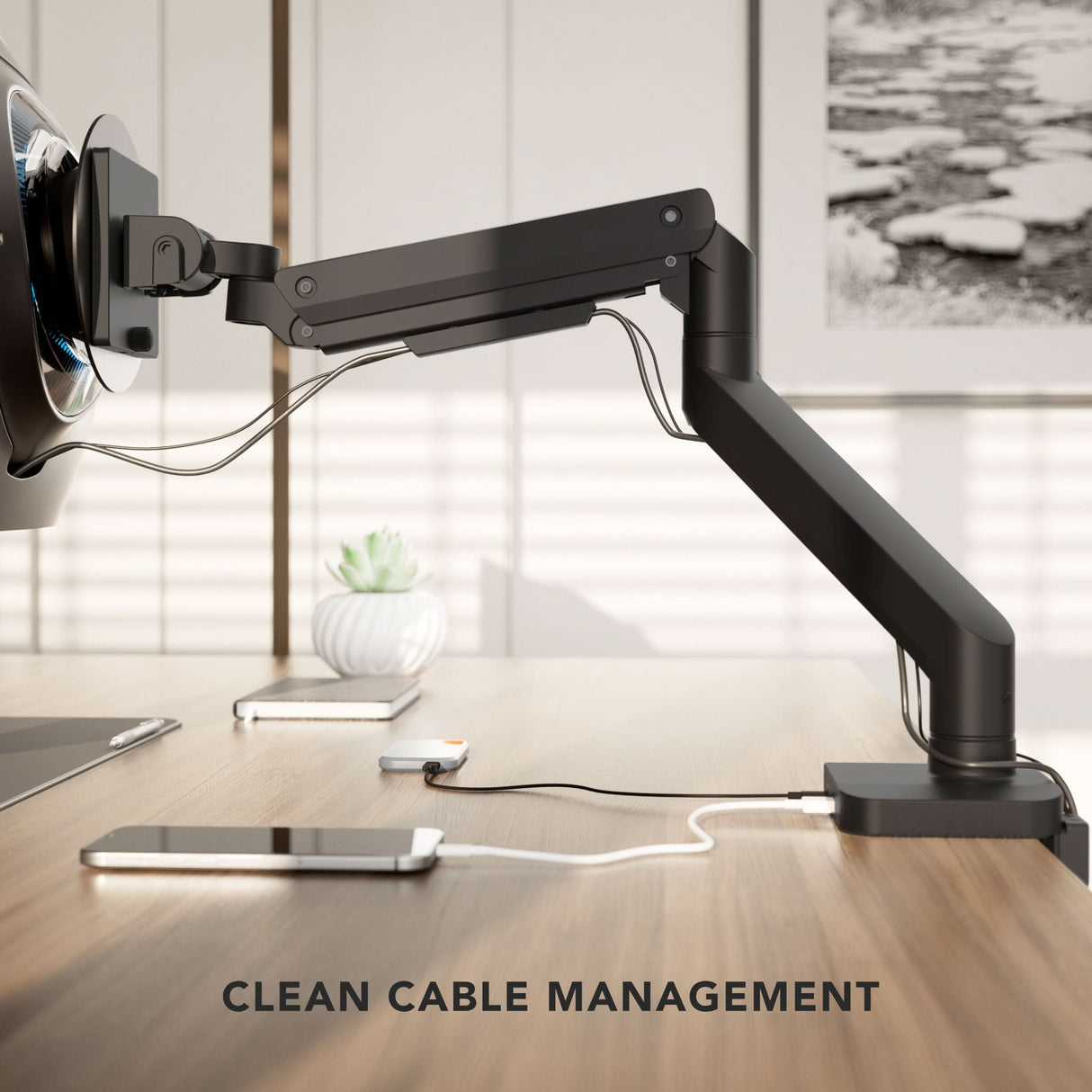 The Mount-It! Heavy-Duty Monitor Mount with Gas Spring Arm holds a screen above a wooden desk, featuring clean cable management. A smartphone and plant are in the background. Text at bottom: CLEAN CABLE MANAGEMENT.