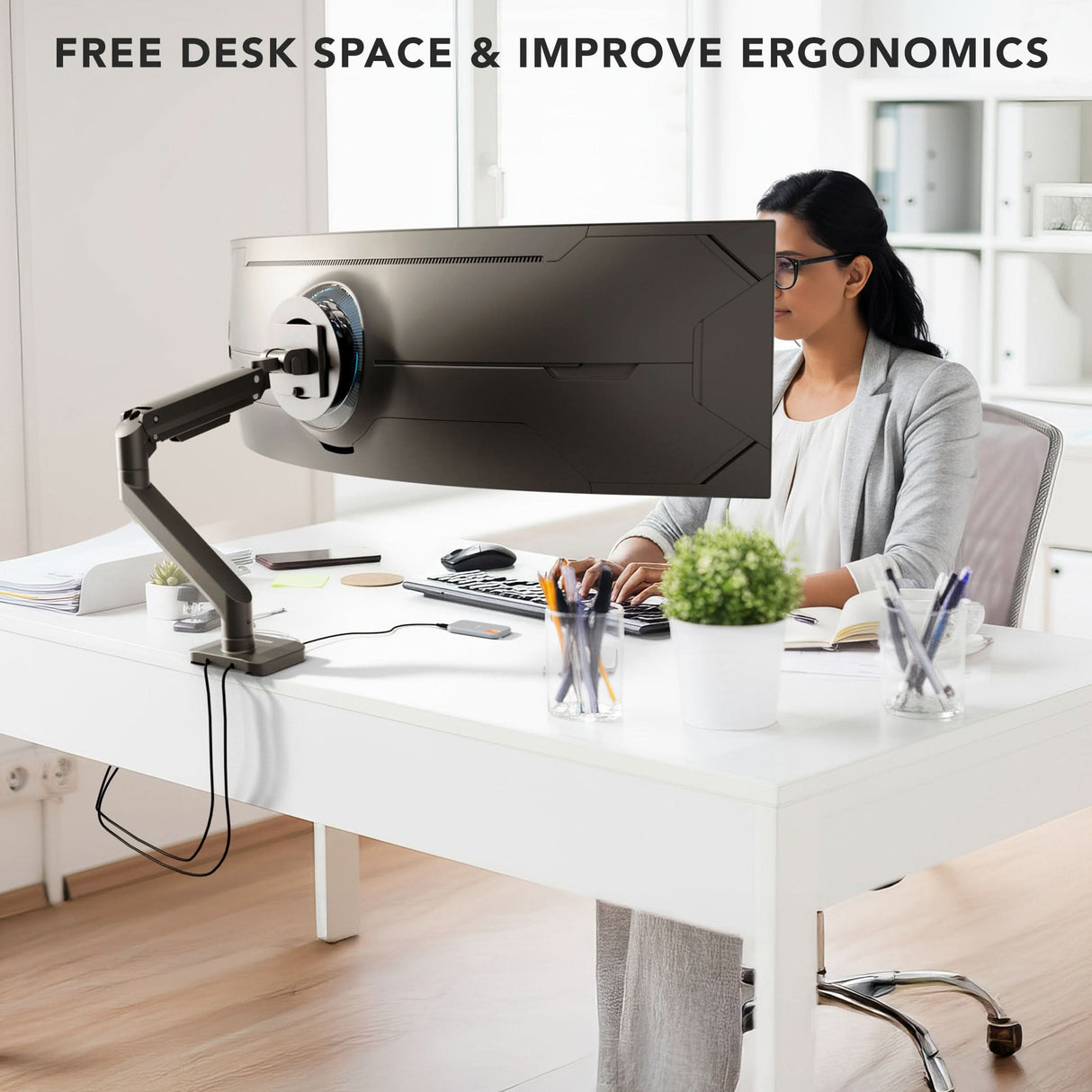 A person uses a large monitor on the Mount-It! Heavy-Duty Monitor Mount with Gas Spring Arm at a white desk in a modern office with shelves, plants, and tidy cables. Text above: FREE DESK SPACE & IMPROVE ERGONOMICS.