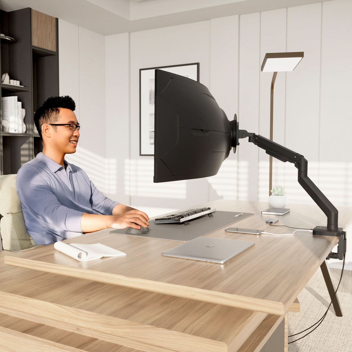 A man sits at a modern desk in a bright office, smiling at a large monitor equipped with a privacy hood and the Mount-It! Heavy-Duty Monitor Mount with Gas Spring Arm. The tidy desk holds a keyboard, mouse, notepad, laptop, and small plant.