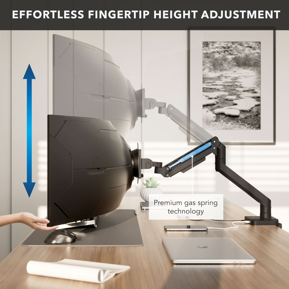 A hand adjusts the Mount-It! Heavy-Duty Monitor Mount with Gas Spring Arm, showing easy fingertip height adjustment and built-in cable management. The workspace also features a laptop, phone, and notebook.