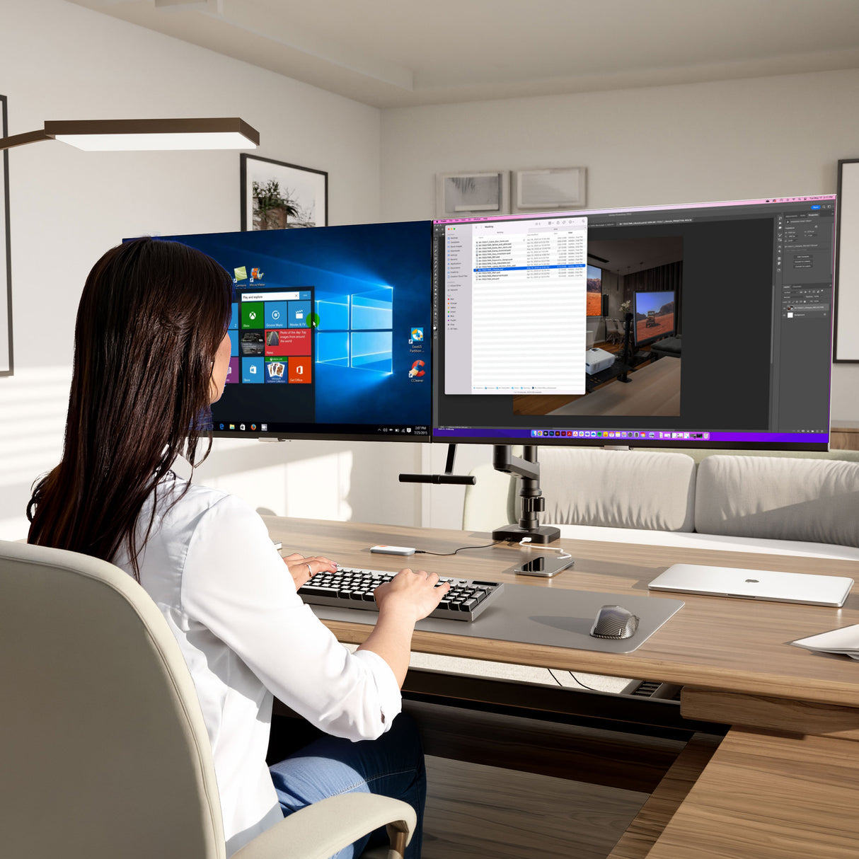 A woman works at a modern desk with the Mount-It! Dual Monitor Mount with Pole and Crossbar, supporting two large monitors—one showing Windows, the other photo editing software—in a bright, minimally decorated office filled with natural light.
