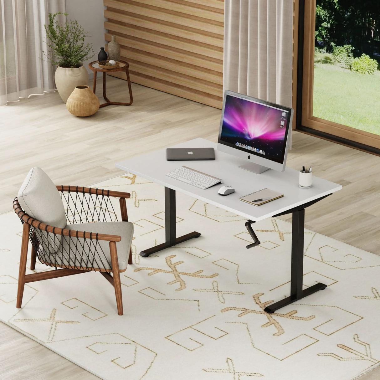 A modern home office showcases the Mount-It! Hand Crank Standing Desk Frame with 48" Tabletop, equipped with a computer, keyboard, mouse, notebook, and pencil holder. A cushioned chair sits on a patterned rug by large windows and indoor plants.