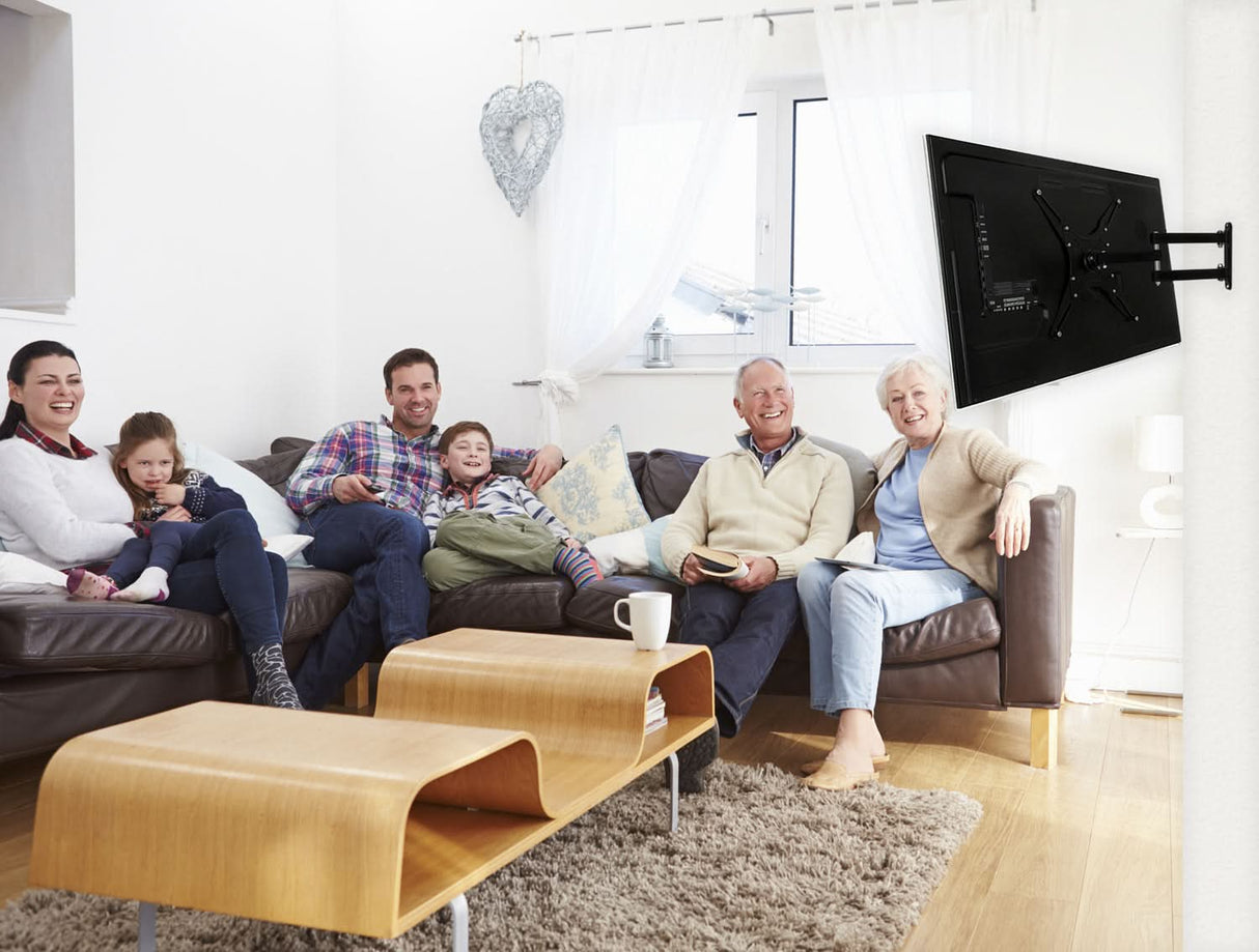 Three generations relax on a sofa in a bright living room, smiling. The TV is mounted with the Mount-It! Full Motion Swivel Bracket TV Wall Mount, and a modern wooden coffee table sits in the foreground.