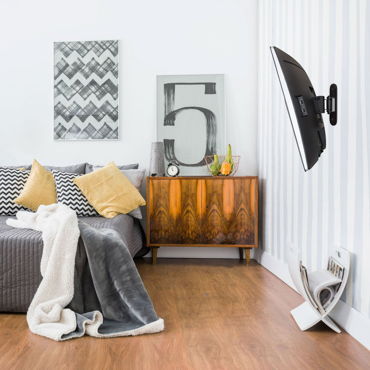 A modern living room with a gray sofa, yellow and gray cushions, a textured blanket, wooden sideboard with decor, abstract artwork, and a Mount-It! Single Stud Tilt and Swivel TV Wall Mount for the wall-mounted TV above the wood floor magazine holder.