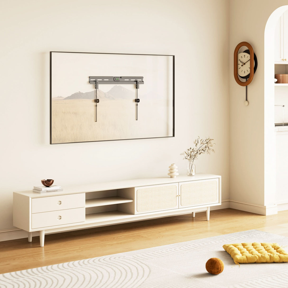A modern minimalist living room with light furniture, a low TV stand, decorative vases, and a yellow cushion. Above the stand, the Mount-It! No Stud TV Wall Mount is framed as art while a vintage clock adds character to the wall.