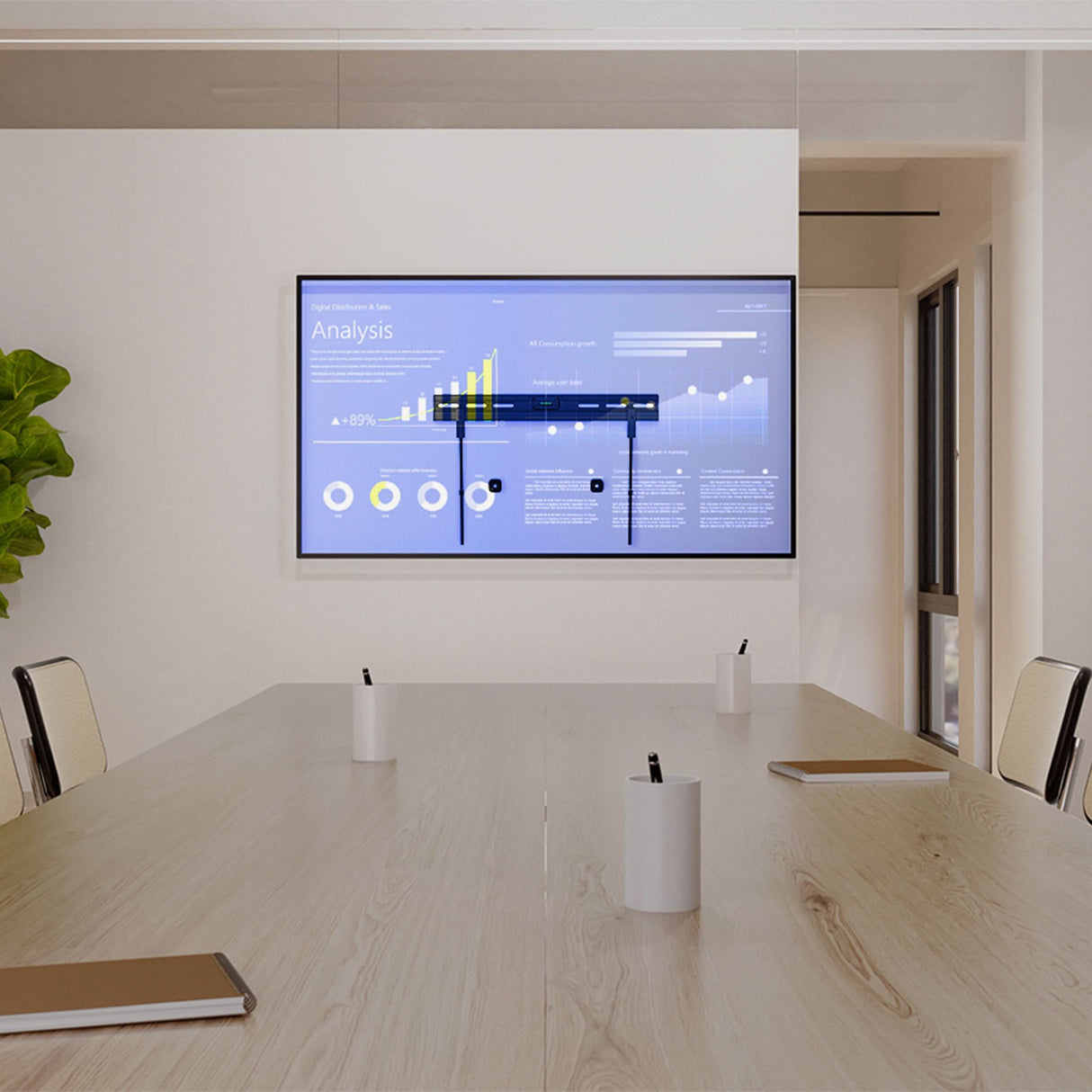 A modern conference room with a wooden table, four chairs, notebooks, pens in holders, and a large TV on the Mount-It! No Stud TV Wall Mount displaying data charts. A green plant sits in the corner.