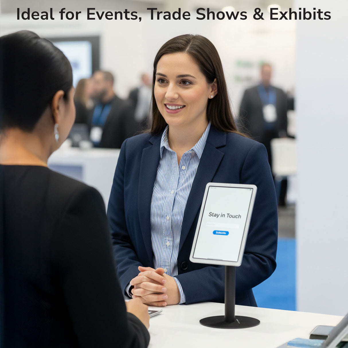 At a trade show booth, a businesswoman chats with a visitor as the Mount-It! Universal Anti-Theft Tablet Countertop Stand displays a “Stay in Touch” message. Text above: Ideal for Events, Trade Shows & Exhibits.