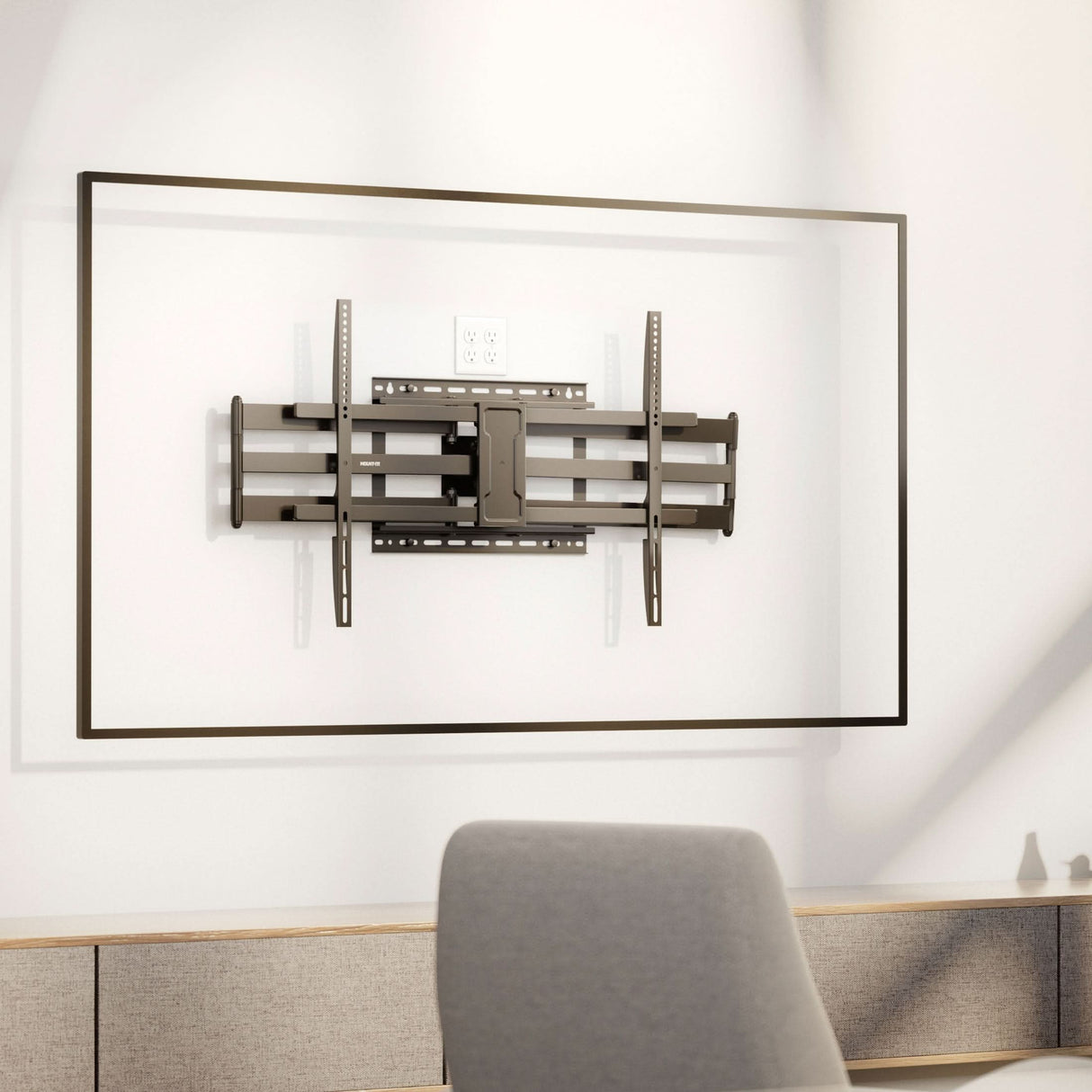 A Mount-It! Full Motion TV Wall Mount with Extra Long Extension is centered in a rectangular frame on a white wall with an outlet above, while a desk and gray chair below create a clean, modern look.