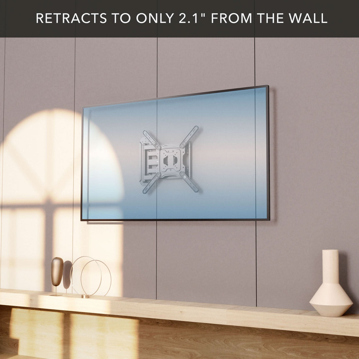 A Mount-It! Full Motion TV Wall Mount with Extra Long Extension supports a flat-screen TV, extending just 2.1 inches from the wall. Sunlight casts shadows above a minimal wooden shelf with decor below the mounted display.