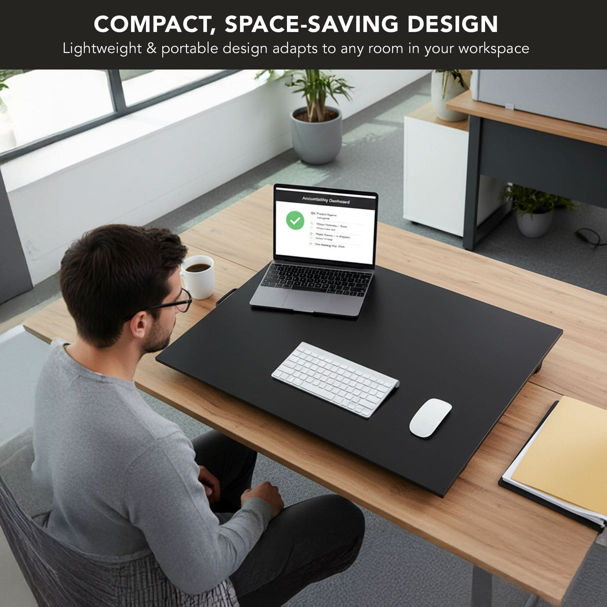 A man works at a Mount-It! Height Adjustable X-Lift Standing Desk Converter with a laptop, keyboard, and mouse on its large black mat. The minimal workspace features ergonomic design and space-saving compactness, highlighted in the text above.