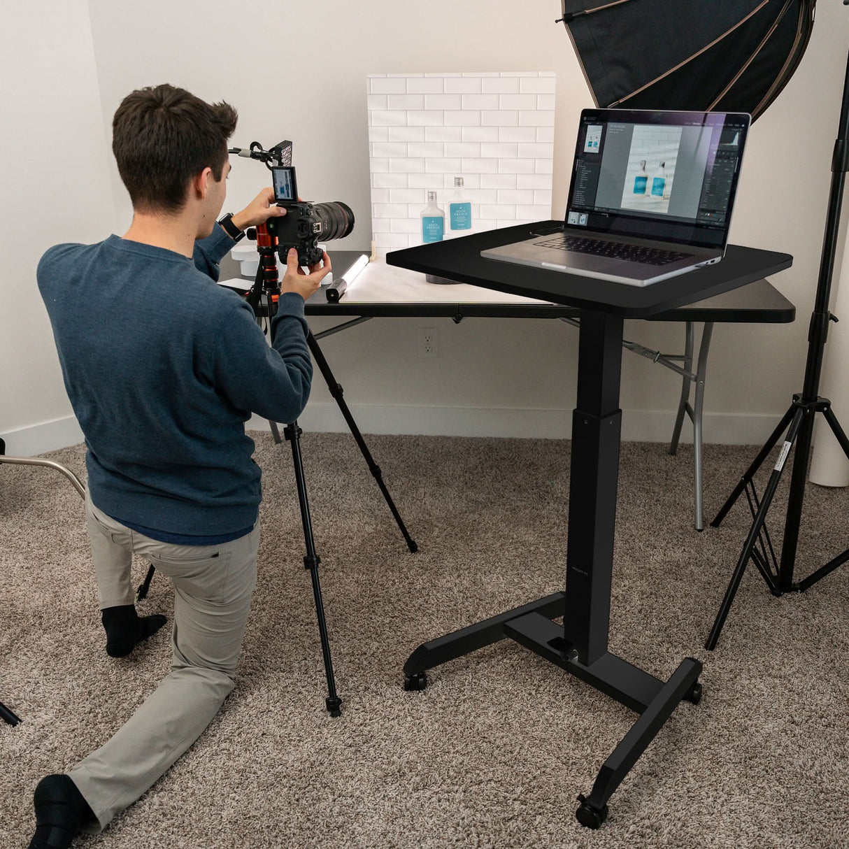 A man kneels on the carpet, photographing products on a table with a camera. Nearby stands the Mount-It! Height Adjustable Rolling Laptop Desk, and photography lighting equipment surrounds the workspace.