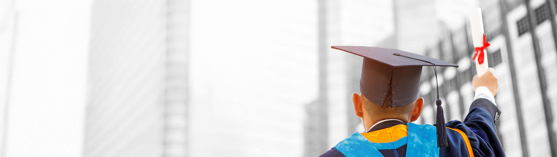 A graduate wearing a cap and gown faces away from the camera, holding up a diploma with a red ribbon in their right hand. The background features tall, modern buildings blurred out, emphasizing the graduate's accomplishment and the moment of celebration in an urban setting. The image captures a sense of achievement and future potential.
