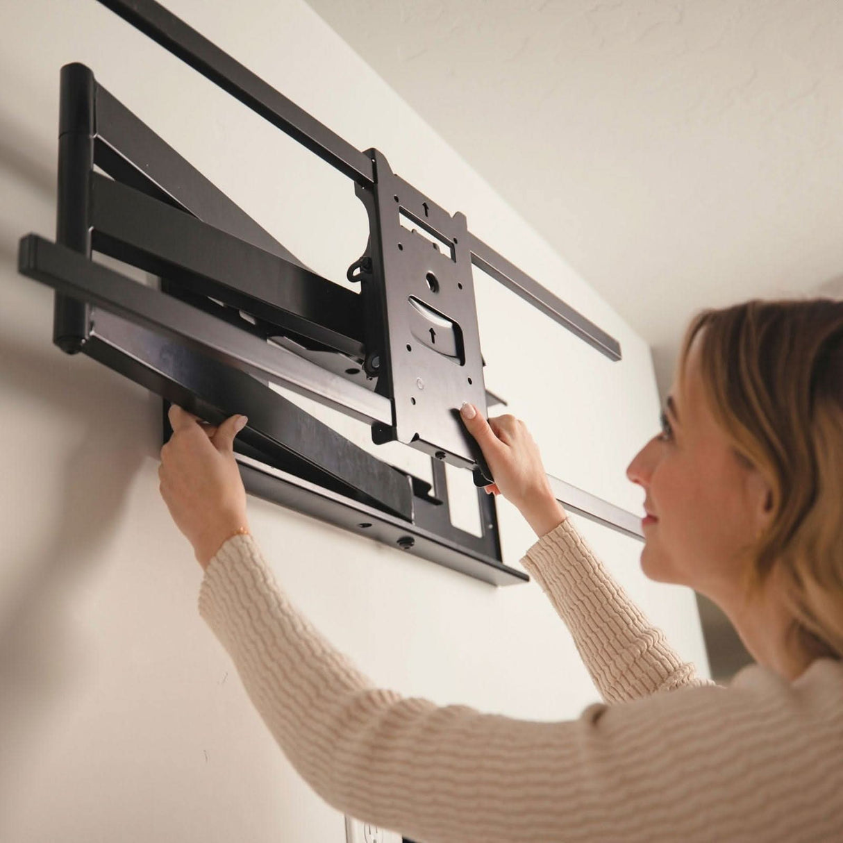 A person with light brown hair in a beige sweater installs the Mount-It! Full Motion TV Wall Mount with Extra Long Extension on a white wall, adjusting the black bracket to support an extra large screen.