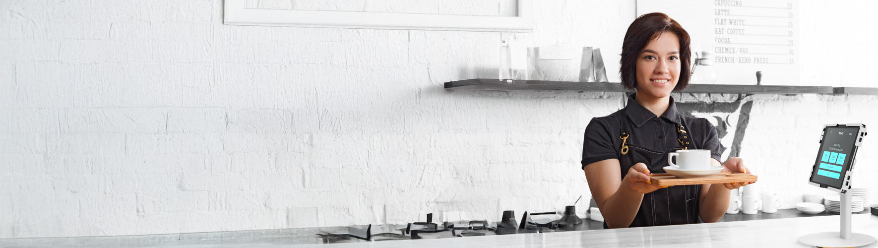 A smiling barista standing behind a clean white countertop in a modern café, holding a tray with a white coffee cup and saucer. The background features a white brick wall with minimalistic decor, including a small shelf with coffee-making tools and a device mounted on the counter displaying a digital screen. The overall atmosphere is bright and inviting.