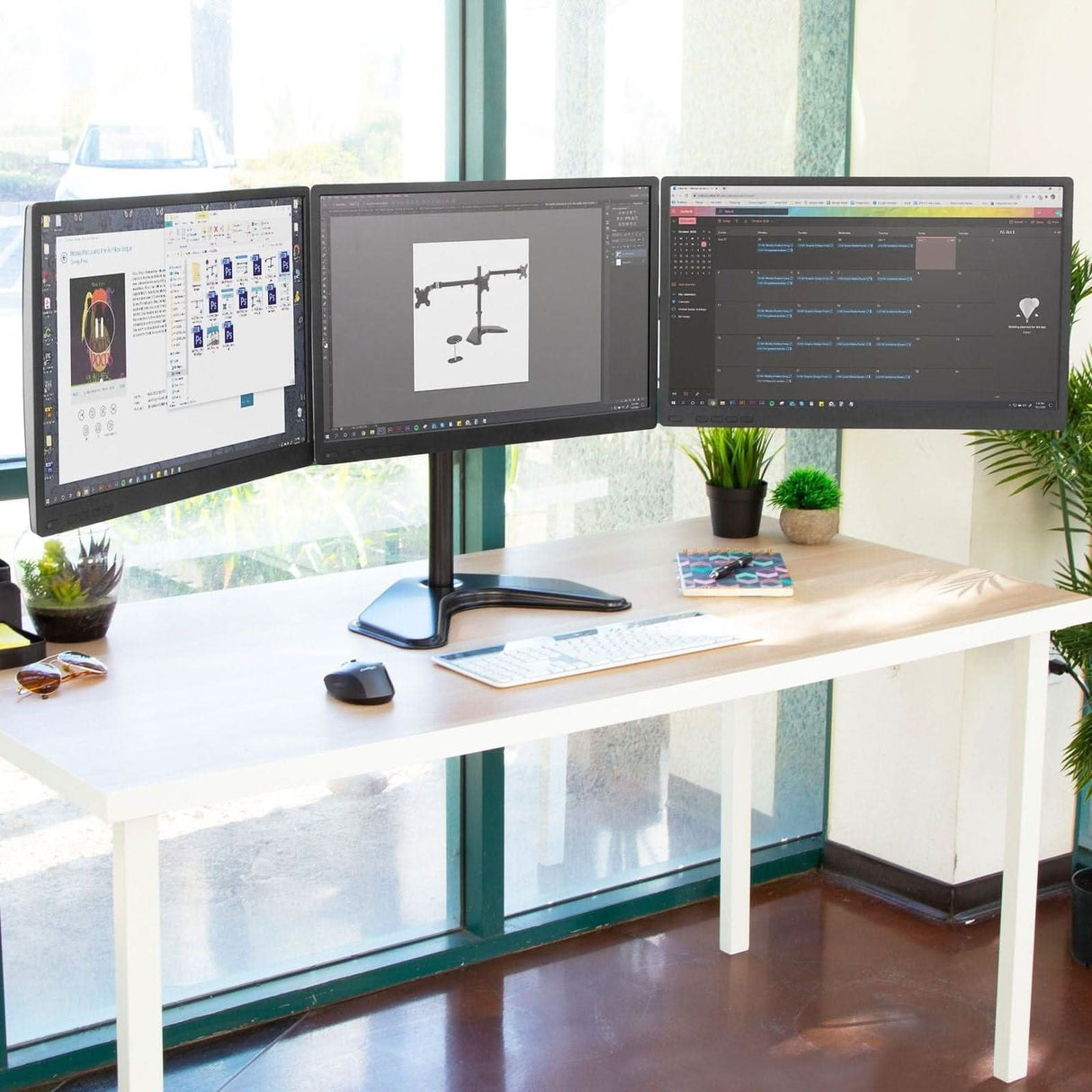 A modern office desk features the Mount-It! Triple Monitor Stand holding three large screens, with a keyboard, mouse, plants, and a magazine nearby. Positioned near large windows, the workspace is bathed in natural light and overlooks greenery.