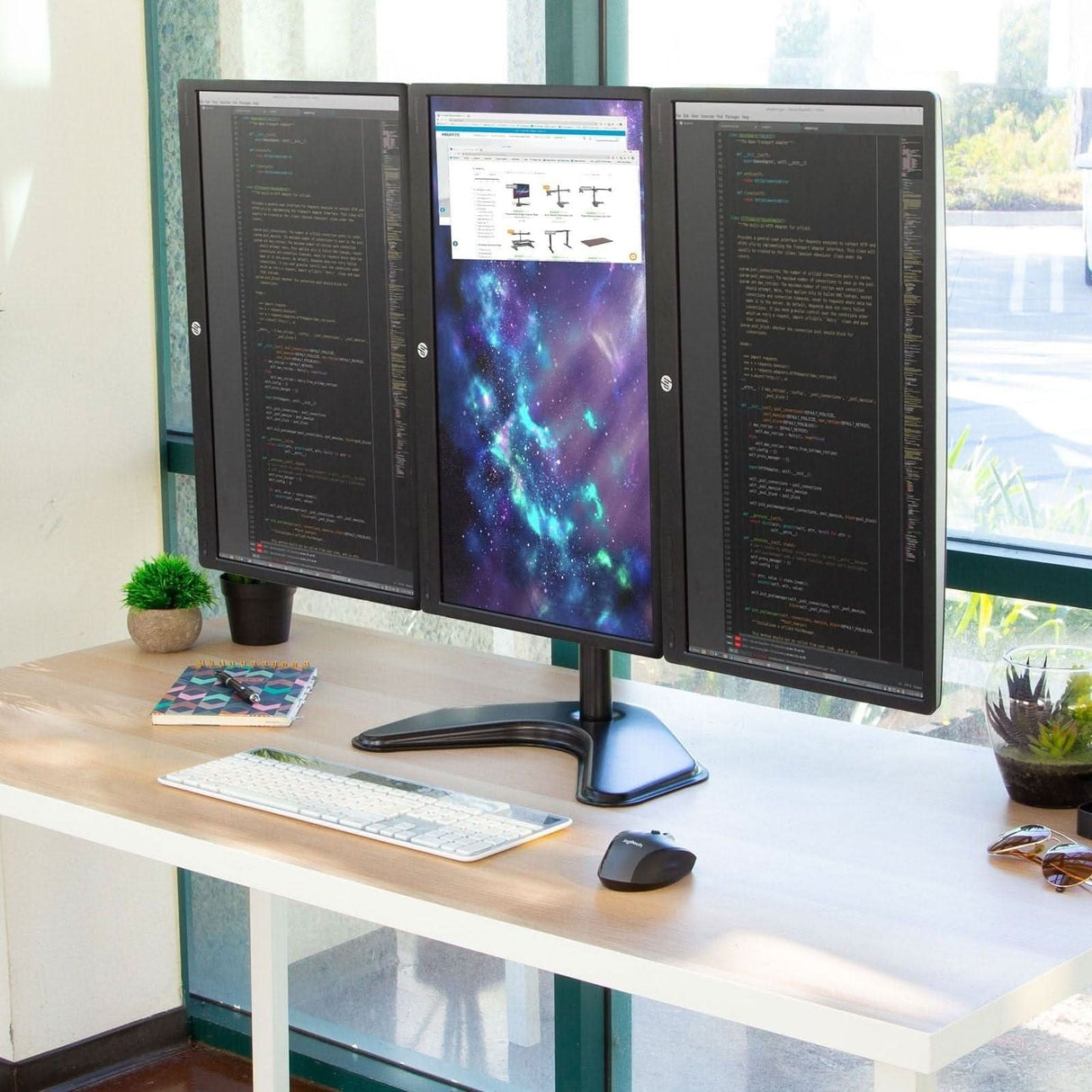 A tidy desk features a Mount-It! Triple Monitor Stand with split dual monitors in vertical alignment displaying code, a cosmic wallpaper, and a web page. The workspace includes a keyboard, mouse, small plants, and sunlight from large windows.