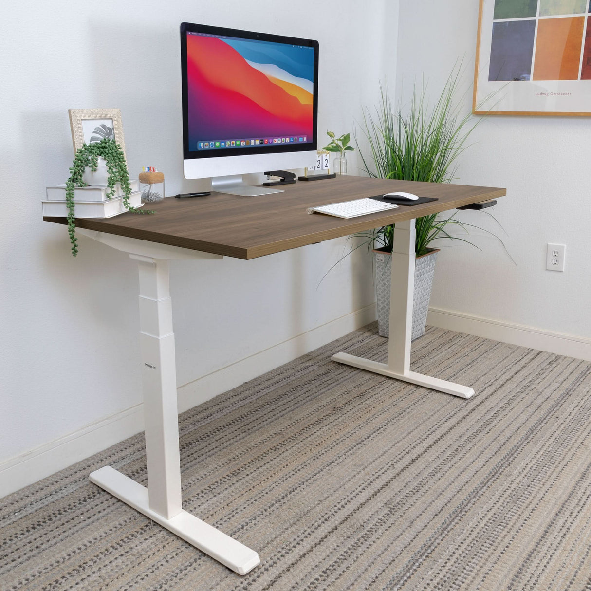 A modern home office setup includes a Mount-It! Ultimate Dual Motor Electric Standing Desk with 55" tabletop, computer, keyboard, mouse, books, small clock, and potted plants on a patterned carpet beside a wall with framed art.