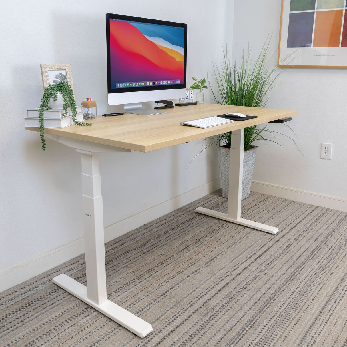 A modern workspace features the Mount-It! Ultimate Dual Motor Electric Standing Desk with 55" tabletop, holding an iMac, keyboard, mouse, small clock, books, framed photo, and potted plant. A large plant and colorful wall art complete the setup.