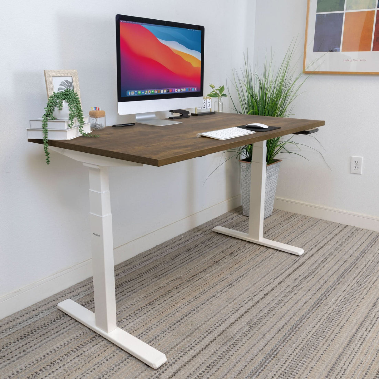 A modern home office features the Mount-It! Ultimate Dual Motor Electric Standing Desk with a 55" tabletop, computer, keyboard, books, plants, and framed art on a light wall—ideal for an ergonomic workspace above the carpeted floor.