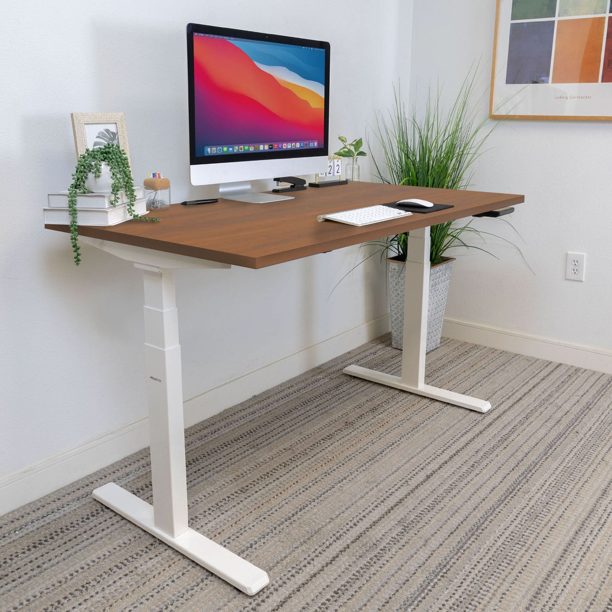 A modern home office features the Mount-It! Ultimate Dual Motor Electric Standing Desk with 55" tabletop, a computer monitor, keyboard, mouse, books, a small plant, tall potted plant in the corner, and artwork above a striped rug.