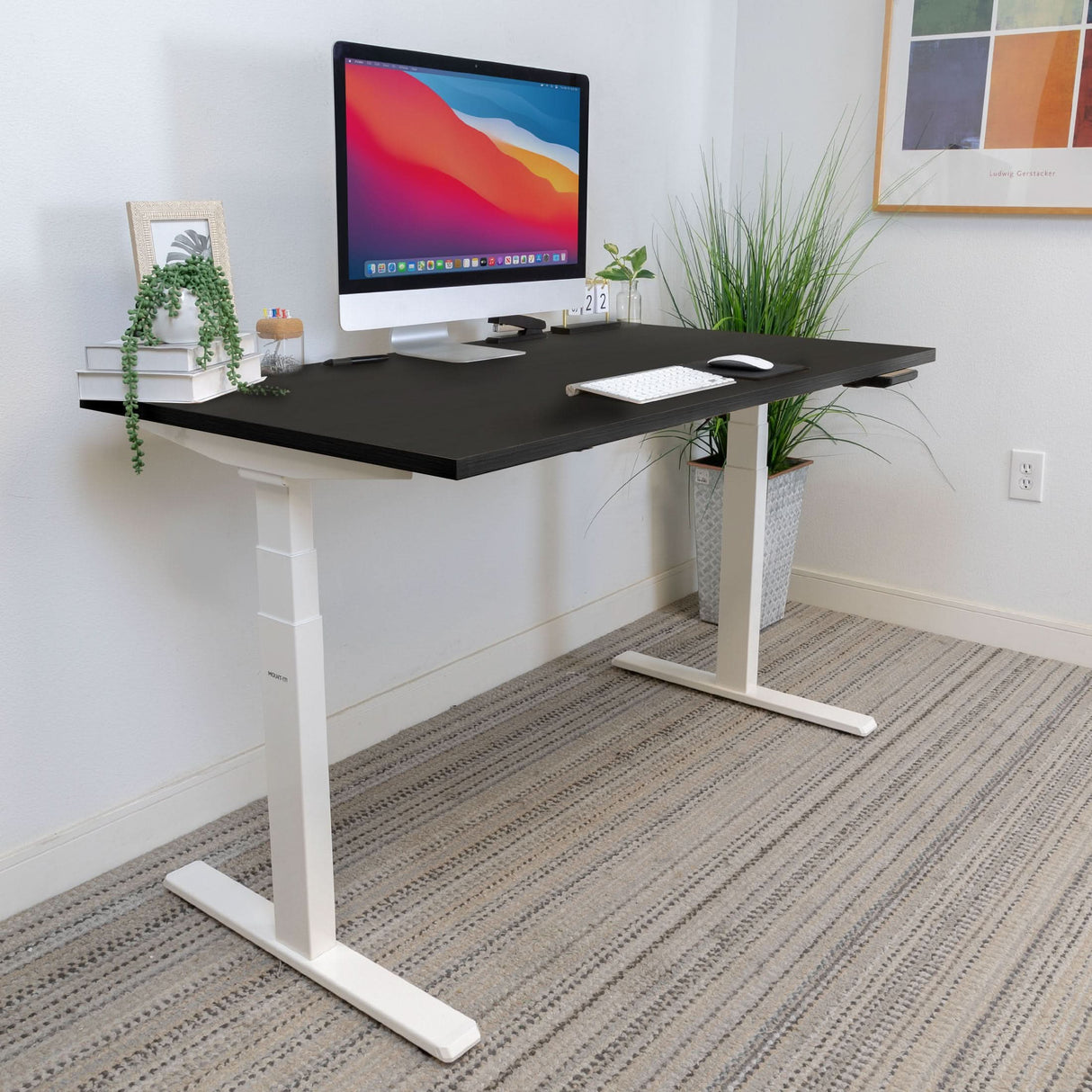 A modern home office showcases the Mount-It! Ultimate Dual Motor Electric Standing Desk with a 55" tabletop in black and white, paired with a computer, accessories, plants, and artwork on a white wall above a striped carpet.