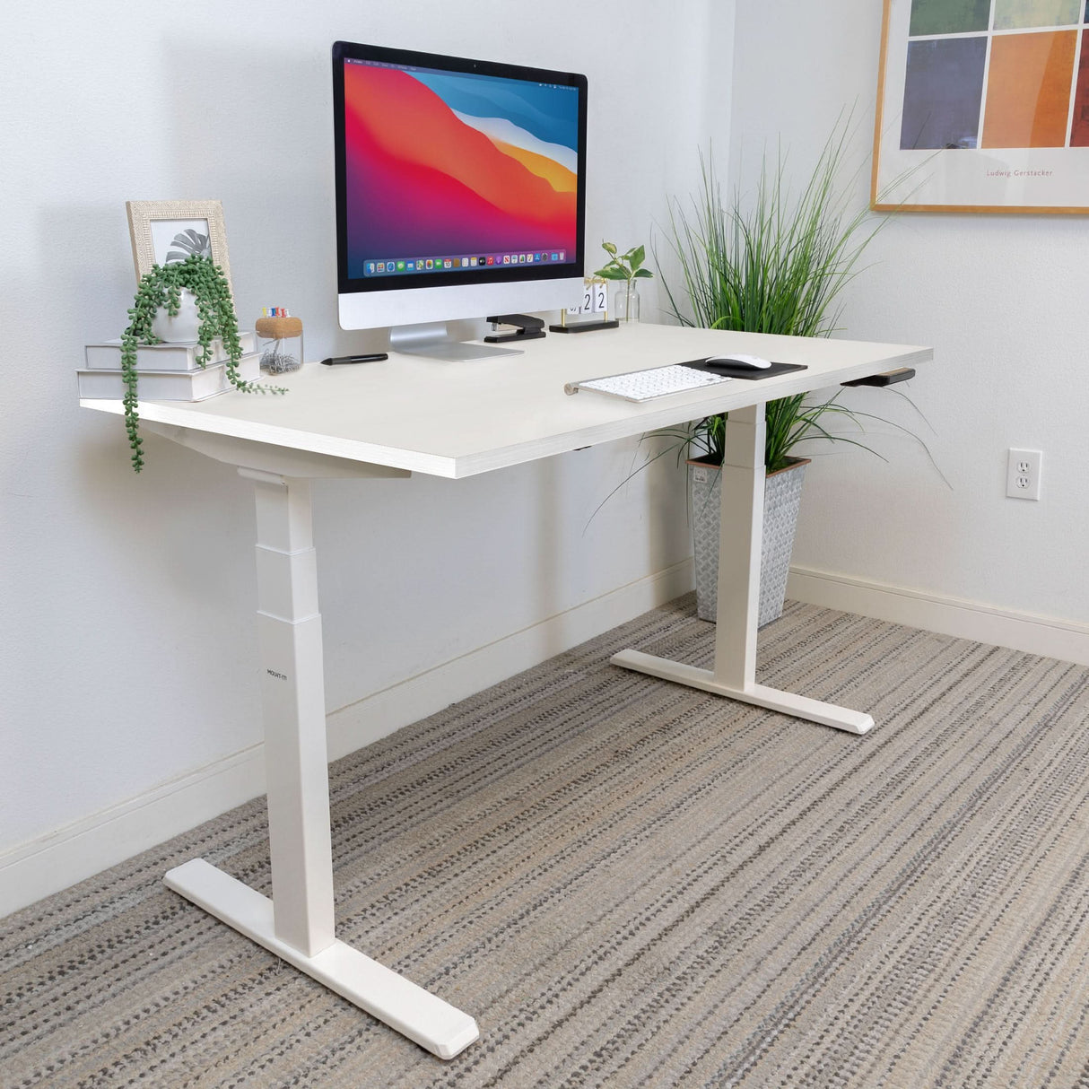 The Mount-It! Ultimate Dual Motor Electric Standing Desk with 55" tabletop features a modern white design, topped with a monitor, keyboard, mouse, potted plant, books, and photo frame. Set by a white wall with artwork and a tall plant for an ergonomic workspace.