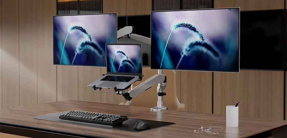 A modern office setup featuring a wooden desk with a dual monitor arm holding two large screens side by side and a laptop mounted below them. The desk also has a keyboard, mouse, clear desk pad, and a small white container holding pens. The background consists of wooden panel walls, creating a clean and organized workspace environment focused on multitasking and productivity.