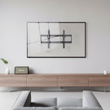 A minimalist living room features a Mount-It! Tilting TV Wall Mount with a slim, art-like frame on a white wall, above a light wood console. A small plant, retro radio, and cup decorate the console; part of a gray couch appears in front.