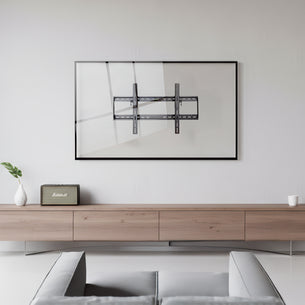 A minimalist living room features a Mount-It! Tilting TV Wall Mount with a slim, art-like frame on a white wall, above a light wood console. A small plant, retro radio, and cup decorate the console; part of a gray couch appears in front.