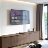 Flat-screen TV mounted on a wall using the Mount-It! Low Profile Tilt TV Wall Mount, showcasing its slim one-inch design that holds TVs from 37 to 80 inches securely with adjustable tilt for glare reduction, installed above a modern wooden cabinet in a bright living room with natural light coming through a window.