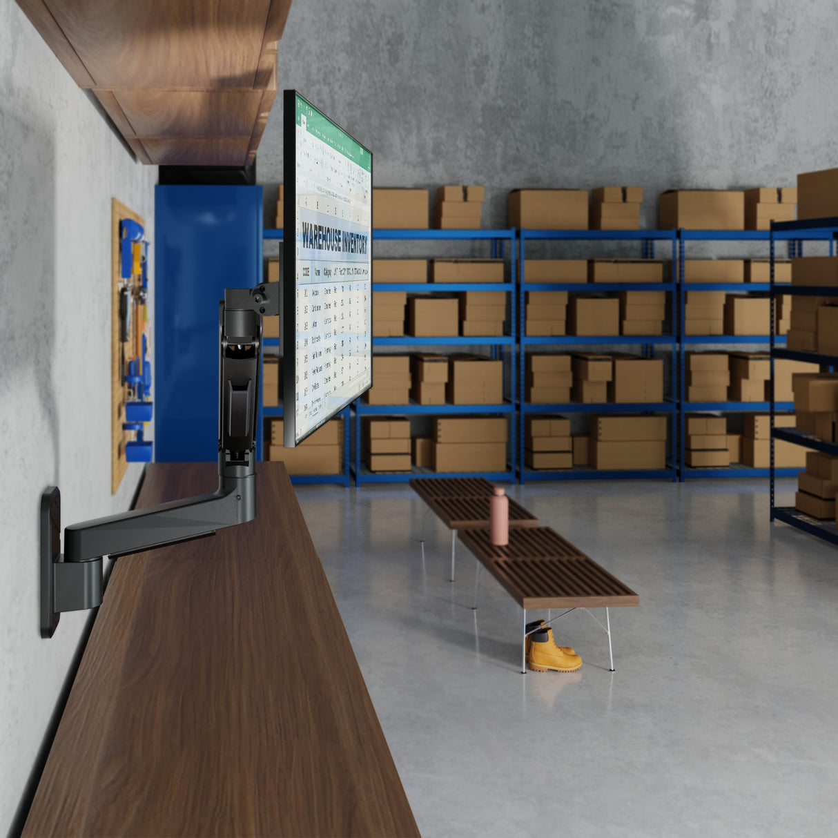 The Mount-It! Heavy-Duty Single Monitor Wall Mount holds a monitor on an adjustable mount attached to a wooden counter in a warehouse with shelves of cardboard boxes, plus a bench and yellow boots—creating an ergonomic workstation.