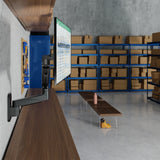 The Mount-It! Heavy-Duty Single Monitor Wall Mount holds a monitor on an adjustable mount attached to a wooden counter in a warehouse with shelves of cardboard boxes, plus a bench and yellow boots—creating an ergonomic workstation.