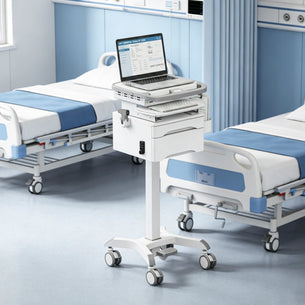 A Mount-It! MedHub Compact Mobile Workstation with Drawers, featuring a laptop and keyboard and equipped with locking casters, stands between two empty hospital beds in a clean, modern room with blue and white decor.
