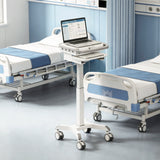 A hospital room with two empty beds and blue-white bedding features the Mount-It! MedHub Motorized Mobile Workstation, equipped with locking casters and an open laptop for patient info—adding a precise, tech-focused clinical atmosphere.