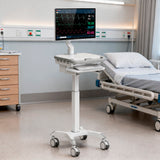 The Mount-It! MedHub Motorized Mobile Workstation, equipped with locking casters, monitor, keyboard, compartments, and tech enclosure, stands beside a hospital bed in an organized patient room.