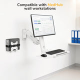A computer monitor, keyboard, and mouse are on a white adjustable mount attached to a wall. The Mount-It! MedHub Mini CPU Holder is also wall-mounted nearby. Blue file folders rest on a shelf. Text reads, Compatible with MedHub wall workstations.