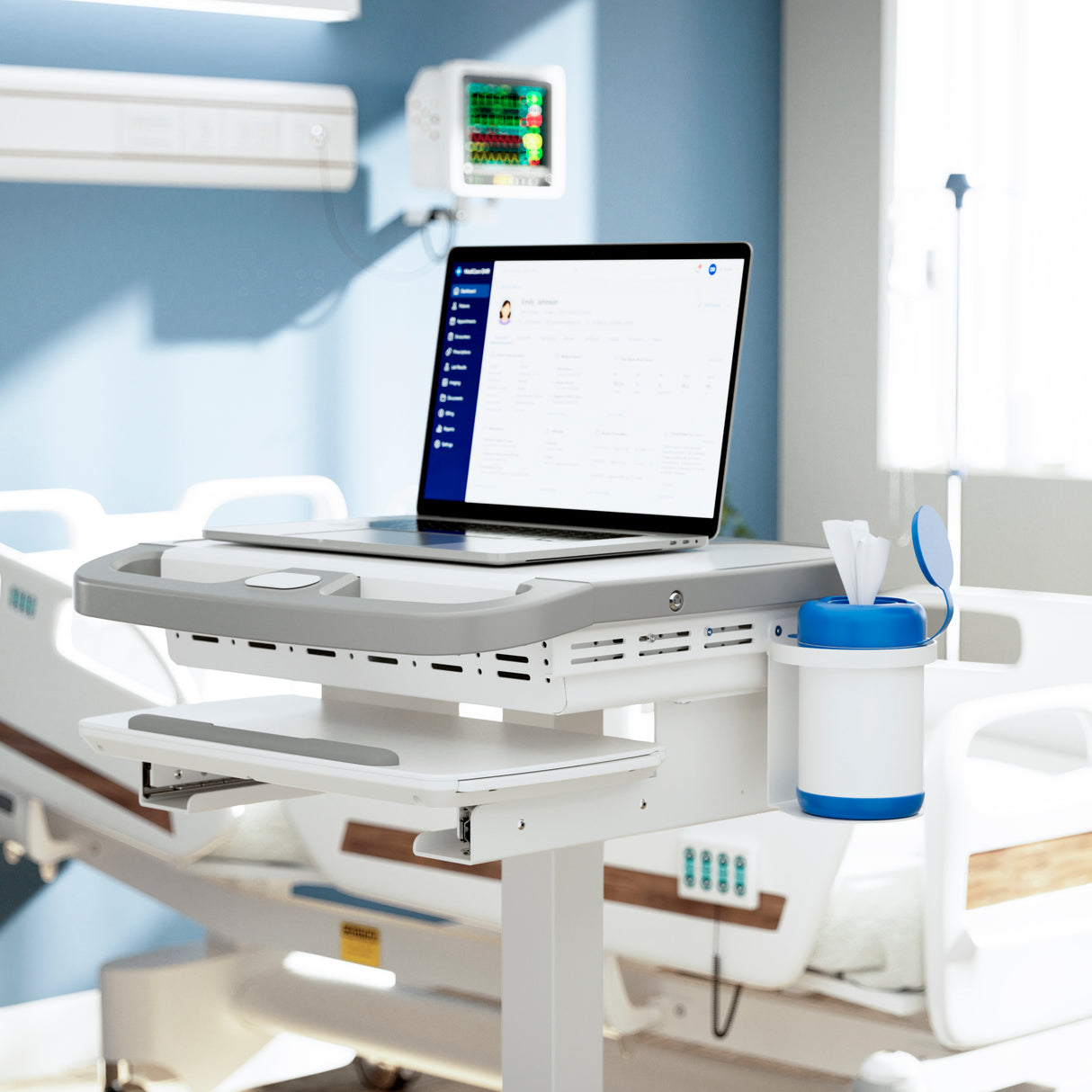 A hospital room with a Mount-It! MedHub Sanitizer Wipes Holder on a mobile workstation next to an open laptop. A hospital bed and monitoring equipment appear in the background, indicating a clinical care setting.