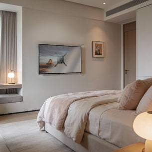 A modern, cozy bedroom with a neatly made bed, soft beige bedding, a Mount-It! Low-Profile TV Wall Mount, a window seat, a small round lamp, minimalist decor, and warm natural light filling the room.
