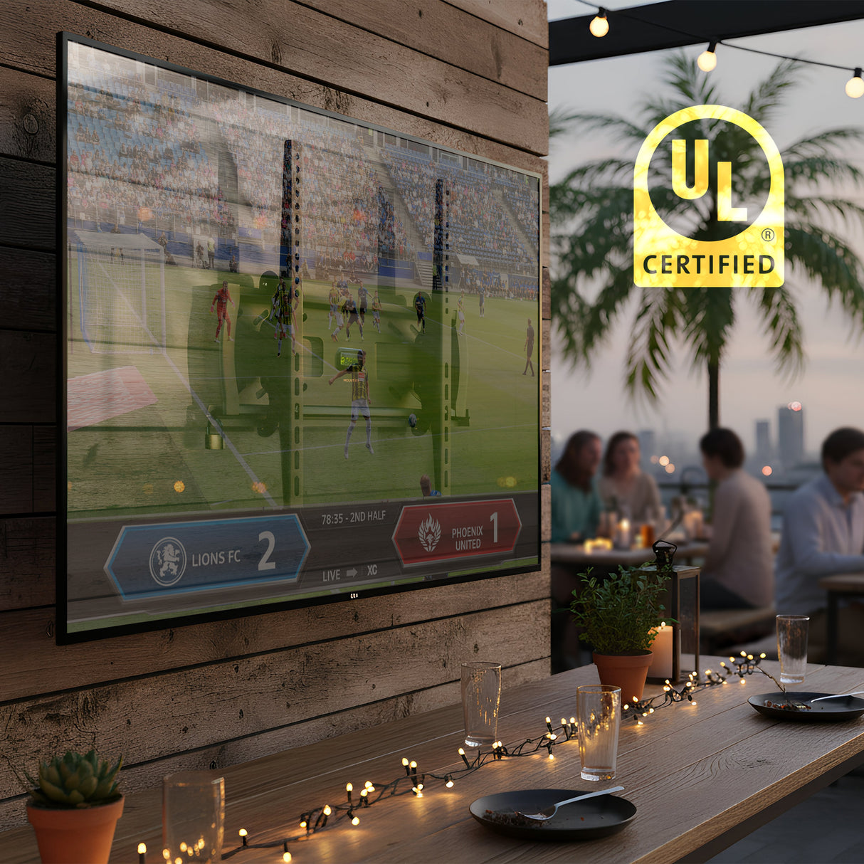 A Mount-It! Locking Anti-Theft TV Wall Mount with VESA 400x400 secures a TV displaying soccer outdoors on a wooden wall as guests dine under string lights, with palm trees, city lights, and the “UL Certified” logo in view.