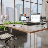 A modern office with a wooden desk, dual monitors on the Mount-It! Full Motion Dual Monitor Desk Mount, plus a keyboard, mouse, chair, and green plants. Large windows showcase city skyscrapers and brighten the space with natural light.