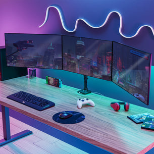 Triple Monitor Mount with Gas Spring Arms and USB Ports holding three curved monitors arranged side by side over a wooden desk with ergonomic keyboard, mouse, gaming controller, headphones, and a handheld gaming console, featuring flexible swivel and tilt adjustments with colorful ambient lighting highlighting a modern workspace.