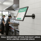 A Mount-It! Point-of-Sale Monitor Stand is wall-mounted in a commercial kitchen, displaying a menu, while two kitchen staff work nearby. Text reads: THE PERFECT SPACE-SAVING POS SOLUTION FOR BUSY COMMERCIAL KITCHENS.