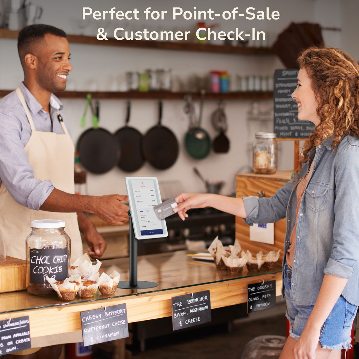 A smiling barista hands a receipt to a customer tapping a card on a touchscreen secured by the Mount-It! Universal Anti-Theft Tablet Countertop Stand, with baked goods and a “Perfect for Point-of-Sale & Customer Check-In” sign nearby.