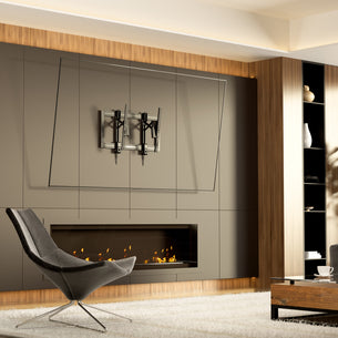 A modern living room features a grey lounge chair, built-in fireplace with wood accents, rug, and an empty TV wall fitted with the Mount-It! Heavy-Duty Advanced Tilt TV Wall Mount and an angled glass guide above the fireplace.