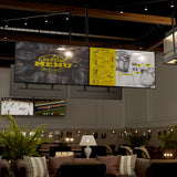 A stylish lounge bar with cozy seating, plants, and wall lamps uses the Mount-It! Dual TV Ceiling Mount with Long Pole for a large digital screen displaying the cocktail menu above and smaller screens below showing sports games.