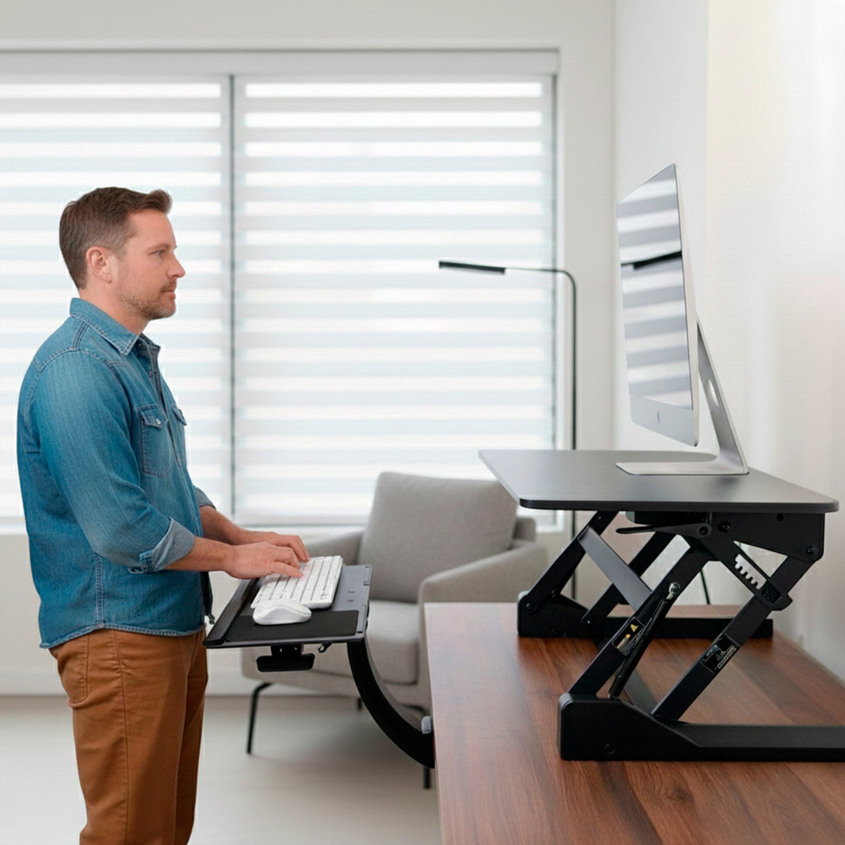 A man types on the Mount-It! Standing Keyboard and Mouse Platform With Ergonomic Wrist Rest Pad at a height-adjustable desk, facing a large monitor in a bright, modern office with a chair and window blinds behind him.