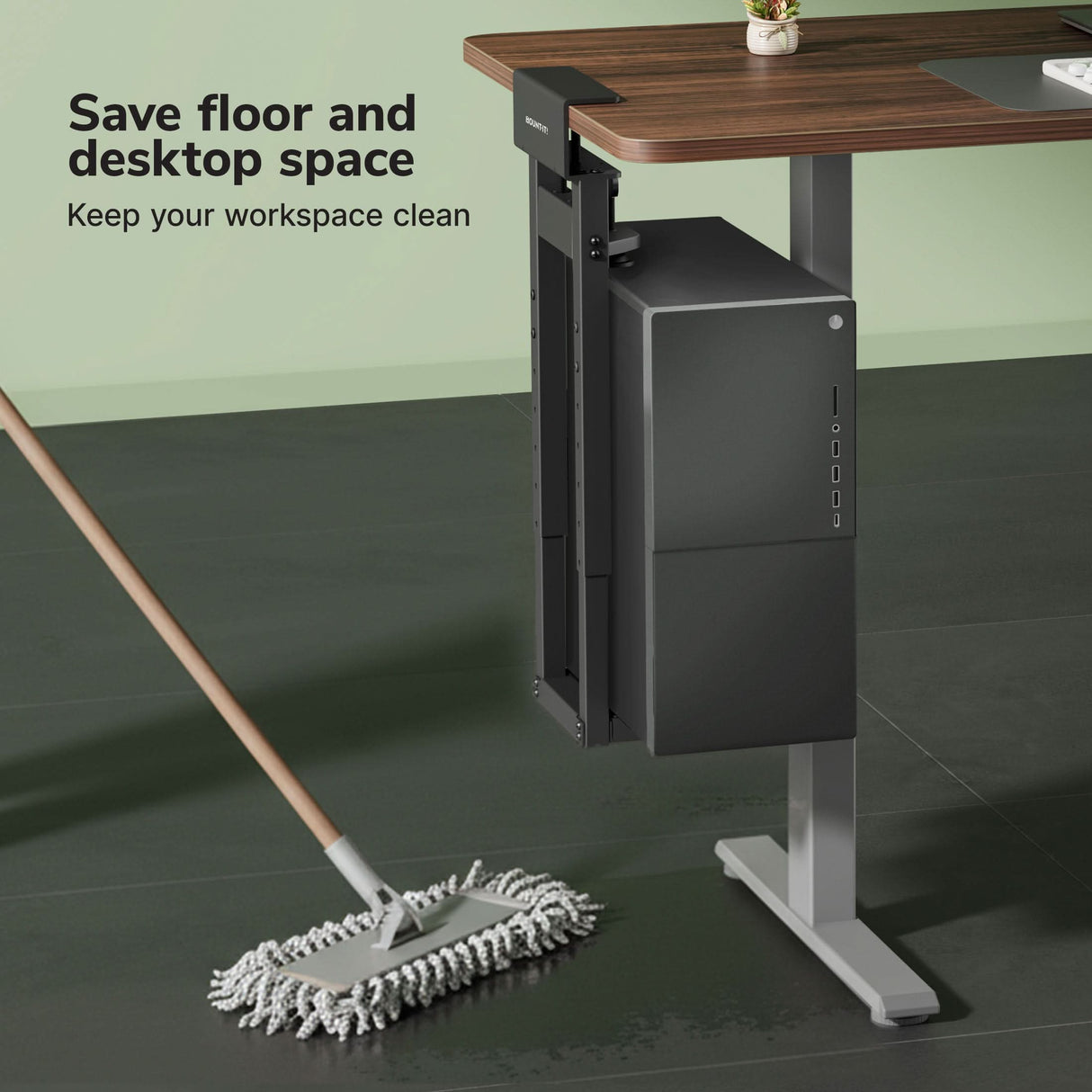 A Mount-It! Heavy-Duty Clamp-On CPU Mount secures your desktop computer under a wooden desk, saving floor and desktop space for a cleaner workspace. A mop wipes the dark floor nearby. Text: Save space. Keep your workspace clean.