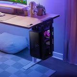 A sleek black computer tower with purple lighting rests in the Mount-It! Heavy-Duty Clamp-On CPU Mount under a white desk by a window. The desk displays a monitor, headphones, and plants bathed in blue and purple ambient lighting.