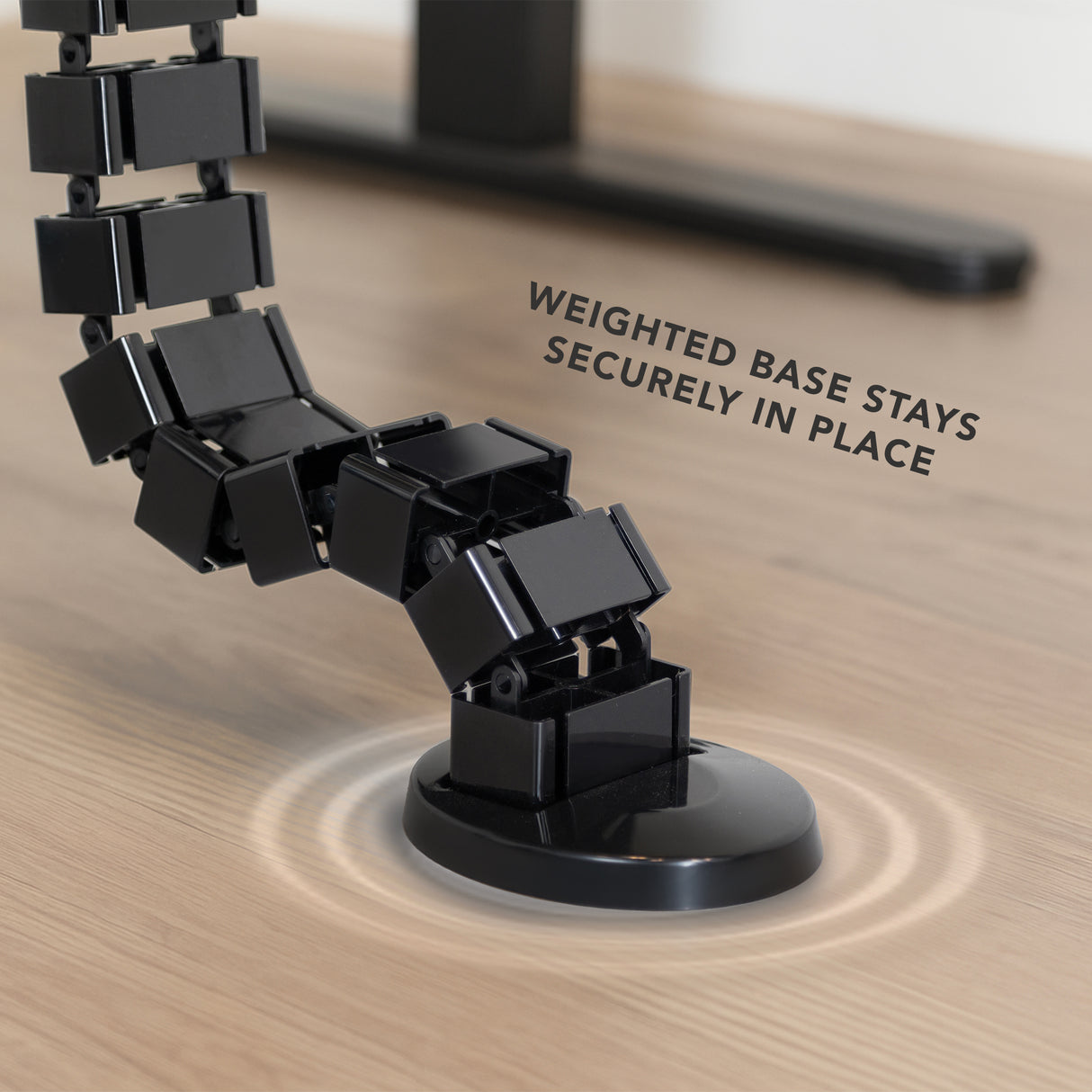 Close-up of the black Cable Management Spine featuring flexible snap-together segments with a weighted base resting securely on a wooden floor, designed to organize and protect cables while adjusting to desk height and providing strain relief.